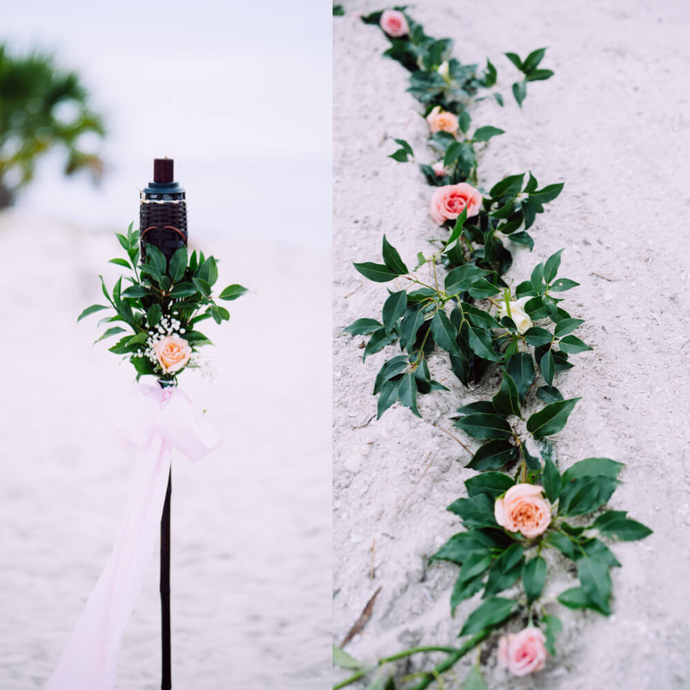 Photo showing decorated tiki torch with fresh roses and greenery and perimeter of a heart in the sand with roses.
